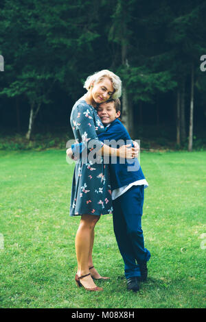 Lächelnde Mutter und Sohn umarmen vor einem Wald auf einer Wiese im Sommer. Stockfoto