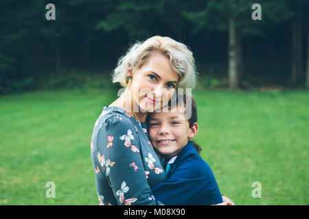 Lächelnde Mutter und Sohn umarmen vor einem Wald auf einer Wiese im Sommer. Stockfoto