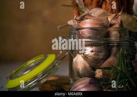 Horizontale Foto von Mehrere Knoblauchzehen. Die Nelken haben dunkle Haut mit violetten Farbton. Gemüse wird in Glas Glas mit Deckel, hat Grüne gask platziert Stockfoto