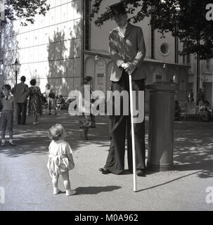 1950 s, historischen, "wer Sie suchen Kid.... ein Kleinkind sieht in Fragen, die mit einem sehr hohen Mann, ein stiltwalker, ein männlicher street Entertainer trägt einen Hut und einen Stock, England, UK. Stockfoto