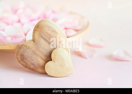 Weiche Bild von zwei Holz- Herzen vor der Platte mit Rosenblättern gefüllt auf rosa Hintergrund Stockfoto