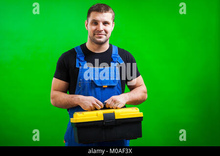 Ein starker Mann Tischler in einem schwarzen T-Shirt und Blauen Bau jumpsuit Lächeln und hält eine Box mit Construction Tools auf seiner Schulter auf der Grünen ist Stockfoto