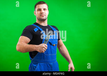 Junger Mann builder Arbeiter in einem schwarzen T-Shirt und Blauen Bau die hält eine neue grau Werkzeug gerade Rohrzange auf einem grünen Hintergrund isoliert Stockfoto