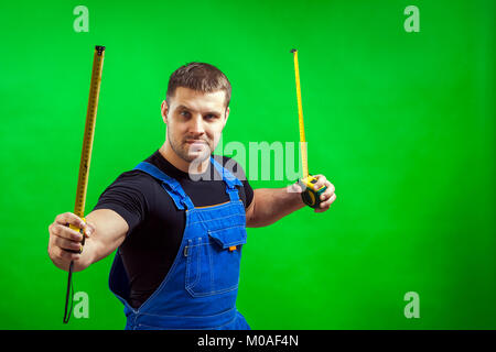 Eine junge männliche Tischler, der ein schwarzes T-Shirt und Blauen Bau jumpsuit hält zwei lange gelbe Band Regel in seinen Händen für die Bauarbeiten für ein Stockfoto
