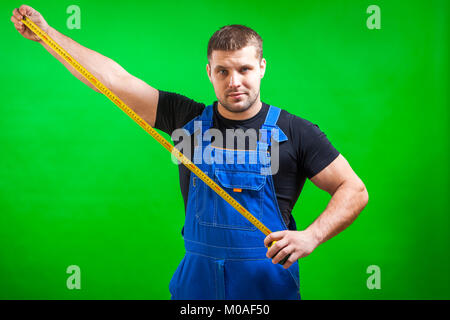 Ein dunkelhaariger Mann Builder in einem schwarzen T-Shirt und Blauen Bau jumpsuit zeigt eine lange gelbe Band Regel, die auf einem Grün isoliert Hintergrund Stockfoto