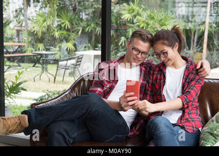Junges Paar in Liebe lächeln & Blick auf Smart Phone. kaukasischer Mann & asiatische Frau sitzen auf dem Sofa zu Hause in der Nähe der Fenster. Freund und Freundin dating im Cafe. Stockfoto