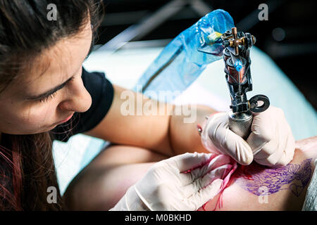 Schöne dunkelhaarige Frau tattoo Master, Tattoo von einem kleinen Vogel mit roten Farben Schreibmaschine für ein Tattoo auf einem weiblichen Oberschenkel, Nahaufnahme, Ansicht von oben Stockfoto