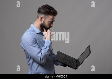 Ernsthafte junge Bartgeier business Mann am Laptop schauen und Denken gegen Grau studio Hintergrund. Stockfoto