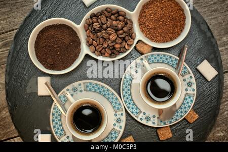 Getränke, Trinkwasser, Energie und Koffein Konzept - zwei Tassen Kaffee, Kaffeebohnen, gemahlenen Kaffee und Instantkaffee auf dunklem Schiefer stand. Stockfoto