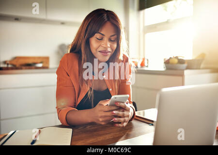 Lächelnden jungen afrikanischen weibliche Unternehmer und das Senden von SMS-Nachrichten, die an einem Notebook arbeitet, während an ihrem Küchentisch sitzen Stockfoto