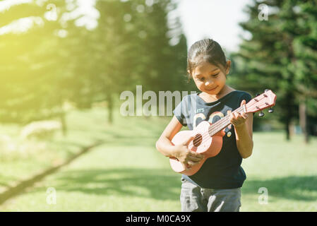 Junge und glückliche asiatischen Mädchen spielt ukelele Gitarre im Park am sonnigen Morgen. Hobbys und Ruhe Konzept Stockfoto