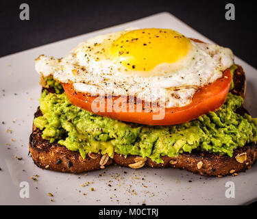 Avocado Toast auf weiße Platte mit Vollkornbrot, Sonnenseite Fired Up Ei und einer in Scheiben geschnittenen Tomaten. Stockfoto