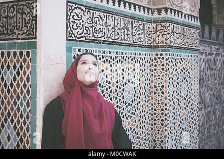 Arabische Frau in traditioneller Kleidung mit roten Kopftuch vor der Wand mit Text aus dem Koran Stockfoto