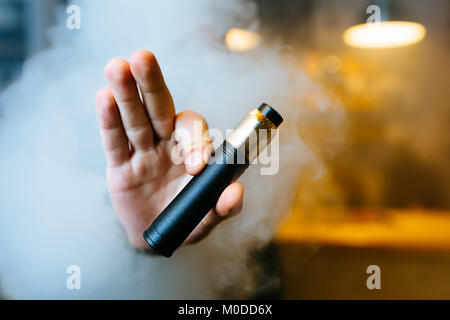 Junge Bart man show vaping Gerät auf seiner ausgestreckten Hand durch eine Wolke aus Dampf. Selektive konzentrieren. Vaping Konzept. Kopieren Sie Platz. Stockfoto