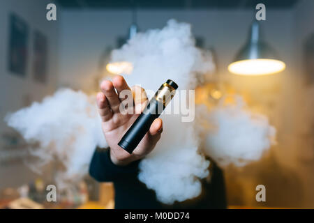 Junge Bart man show vaping Gerät auf seiner ausgestreckten Hand durch eine Wolke aus Dampf. Selektive konzentrieren. Vaping Konzept. Kopieren Sie Platz. Stockfoto