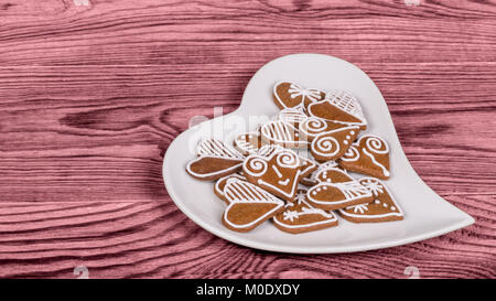 Haufen verzierten Herzen aus Lebkuchen auf weißen herzförmigen Platte. Valentinstag Hintergrund mit Symbole der Liebe und Glück auf rosa-farbige Holz. Stockfoto