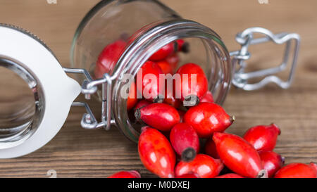 Verschüttete rote Hagebutten auf Holz Tisch. Nahaufnahme der Lüge Glas Glas und Arzneimitteln, Früchte von Wild Rose auf der braunen Hintergrund. Stockfoto