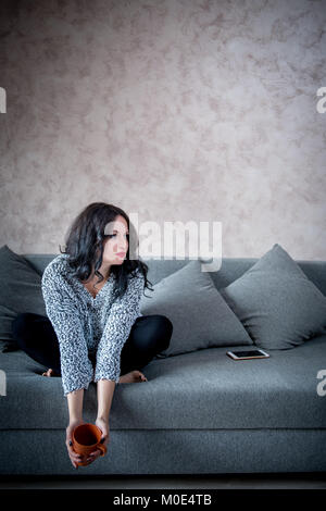 Junge Erwachsene brünette Frau sitzt auf einem Sofa zu Hause mit Handy, besinnlich und nachdenklich Stockfoto