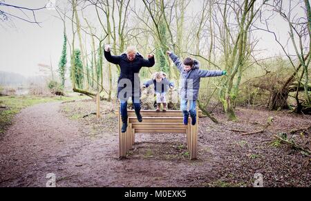 Mutter und zwei Kinder springen von einer Holztreppe im Wald, Kingsbury, Warwickshire, England, Großbritannien Stockfoto