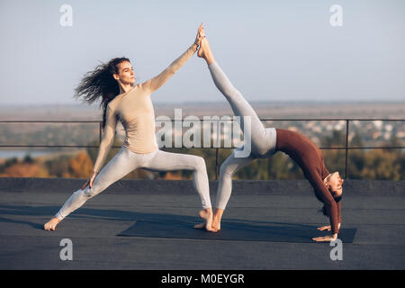 Zwei schöne Frauen in acroyoga beschäftigt, einander helfen, im Freien. Eine Turnerin in der Pose der Krieger, unterstützt einen Anderen, der in der Pose des chakrasana, Stockfoto