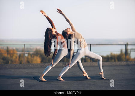 Mädchen synchron durchführen pose Krieger mit erhobener Hand, auf dem Dach im Freien. Schöne Yoga Asana mit Elementen der Akrobatik. Gesundes Leben Stockfoto