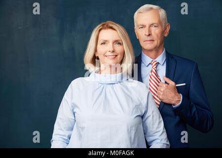 Studio Portrait von Senior Business Leute an dunklen Hintergrund stehen. Wunderschöne reife Frau in T-Shirt und älterer Mann mit Anzug beim Stehen Stockfoto