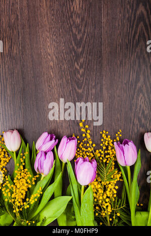 Strauß Tulpen und Mimosa in einer gelben Vase auf einem hölzernen Hintergrund. Stillleben mit Blumen in den Urlaub. Stockfoto