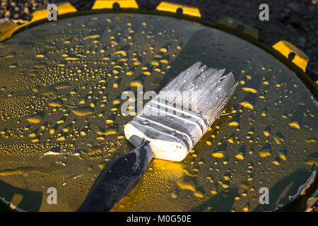 Ansicht von oben in einem unordentlichen Pinsel gehalten auf dem Deckel einer Farbe Eimer auf dem felsigen Boden und viel Regen Wassertropfen auf es am Morgen Stockfoto
