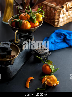 Frische ganze Clementinen mit grünen Blättern in vintage Maßstab und eine geschälte Clementine auf einem Schwarzen Tisch mit einem blauen Tuch und mehrere Gewichte Stockfoto