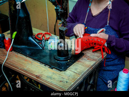 Weibliche Hand eines Schuhmachers näht das Schloss in roten Stiefeln. Auf der Nähmaschine sind die Gewinde und Schere. Stockfoto