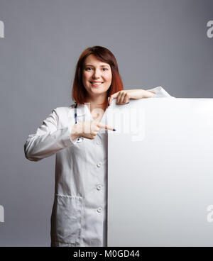 Bild von lächelnden Frau Doktor in weißem Gewand und mit phonendoscope Punkte Finger auf leeren Blatt Pape Stockfoto