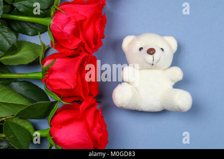 Weißer Teddybär von Rosen auf einem grauen Tabelle umgeben. Vorlage für den 8. März, Muttertag, Valentinstag Stockfoto