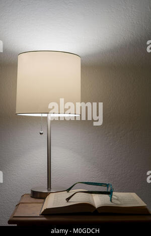 Ein paar gebrochene reader Gläser auf ein altes Buch öffnen ruhend, unter einem Nachttisch Lampe, gegen weiße Wand, was auf eine ältere eine Pause Stockfoto