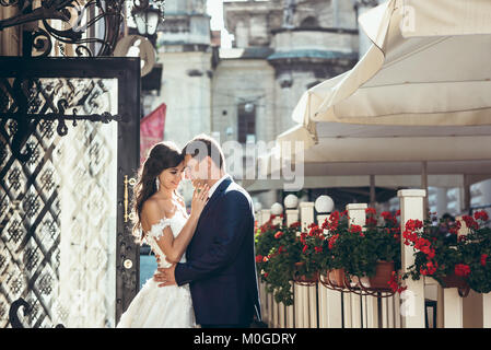 Horizontale Hochzeit Portrait der glücklich umarmte Jungvermählten im sonnigen Straße. Die Braut ist Streicheln der Wange des gutaussehenden Bräutigam. Stockfoto