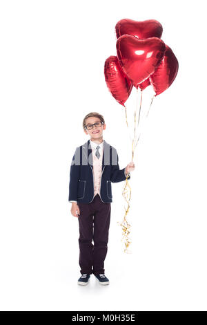 Cute little boy in Anzug und Brille mit roten herzförmigen Luftballons und lächelnd an Kamera isoliert auf weißem Stockfoto