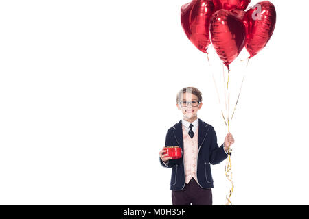 Cute little boy in Anzug und Brille mit roten herzförmigen Luftballons und Geschenkbox isoliert auf weißem Stockfoto