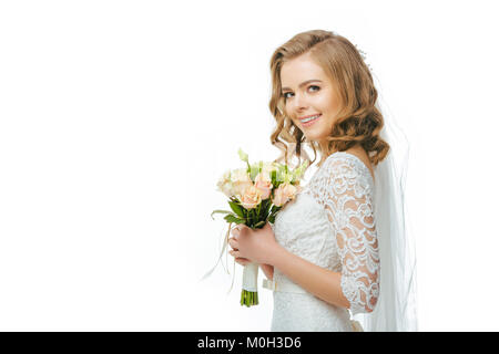 Seitenansicht der lächelnden Braut im Brautkleid und Schleier mit Blumenstrauß isoliert auf weißem Stockfoto