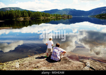 Vater und Sohn sitzen auf einem Felsen am schönen See mit einem Sky Reflexion und Angeln versuchen. Norwegen. Stockfoto