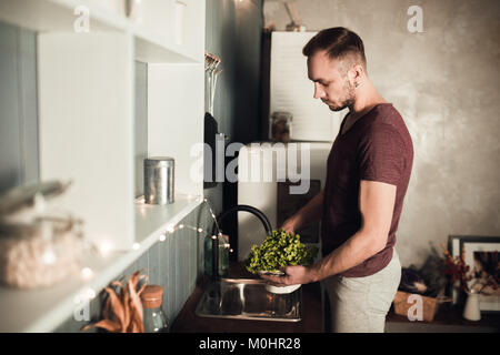 Portrait von lächelnden Mann. Kerl in Sport Home waschen Salat in der Küche. Rustikal Farbe lifestyle Foto von Jungen in loft Interieur. Stockfoto