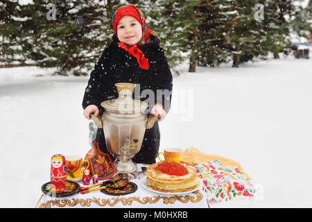 Kind, Mädchen in einem Pelzmantel und in einen Schal im russischen Stil mit einem großen Samovar in den Händen der Pfannkuchen mit roter Kaviar Stockfoto