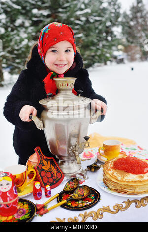 Kind, Mädchen in einem Pelzmantel und in einen Schal im russischen Stil mit einem großen Samovar in den Händen der Pfannkuchen mit roter Kaviar Stockfoto