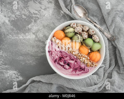 Gesundes und Leckeres Essen - Berry Smoothie und Müsli mit Nüssen und Sesam, Stücke von Kiwi und Persimmon in eine weiße Platte auf eine stylische, graue Lasche Stockfoto