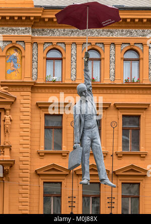 Die Umbrella Man von Prag, gegen ein orangefarbenes Gebäude gesehen, eine moderne Kunst Statue eines Mannes hängt von einem Dach hoch über einem Prager Straße ausgesetzt Stockfoto