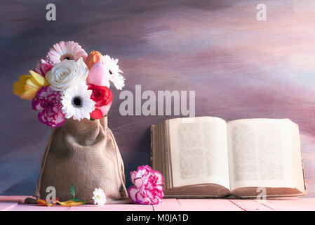 Schönen Blumenstrauß aus Rosen, Gerbera, Tulpen und Nelken auf einem rosa Tabelle und ein antikes Buch öffnen gegen einen lila Holz- Hintergrund lehnte. Stockfoto