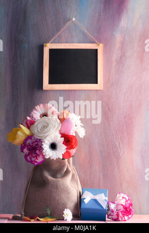 Blumenstrauß aus Rosen, Gerbera, Tulpen, Nelken, in einem Jute Sack, von Blumen und ein Geschenk und eine leere Tafel im Hintergrund umgeben. Stockfoto