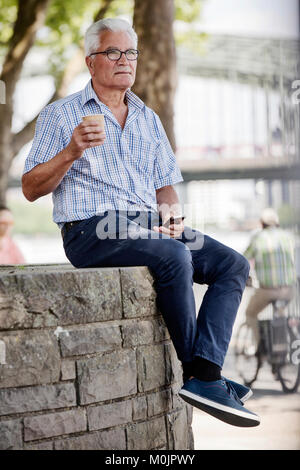 Senior sitzt mit dem Smartphone und einem Kaffee an einer Wand im Kölner Rheinufer Promenade, Köln, Nordrhein-Westfalen zu gehen Stockfoto