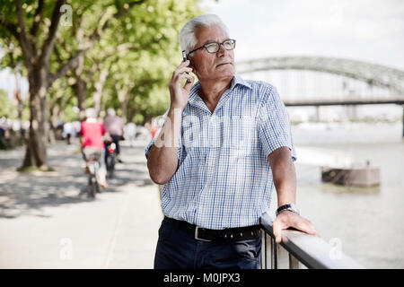 Älterer grauhaariger Anrufe mit seinem Smartphone am Kölner Rheinufer Promenade vor der Hohenzollernbrücke, Köln Stockfoto