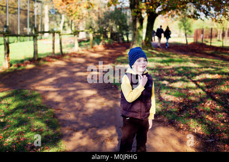Ein lächelnder kleiner Junge in warmer Kleidung ist bereit, in einem goldenen Herbstpark in Großbritannien Spaß zu haben. Herbstlicht, Freiheit, Glück, Kindheit. Stockfoto