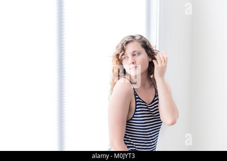 Seite Nahaufnahme Porträt einer jungen Frau im weißen, hellen Zimmer des Hauses, zu Hause sitzen, Appartement suchen durch sonnige große weiße Fenster mit Jalousien Stockfoto