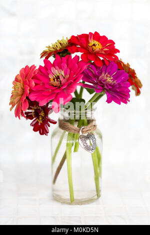 Eine Vielzahl von farbigen Zinnia Blumen in einem einfachen Glas Vase mit einem dekorativen Herzanhänger mit einem Jute Schnur gehalten Stockfoto